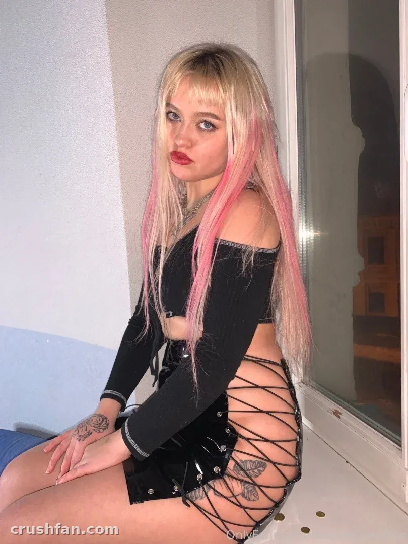 Creator with pink-streaked blonde hair in a dark outfit seated by a window indoors