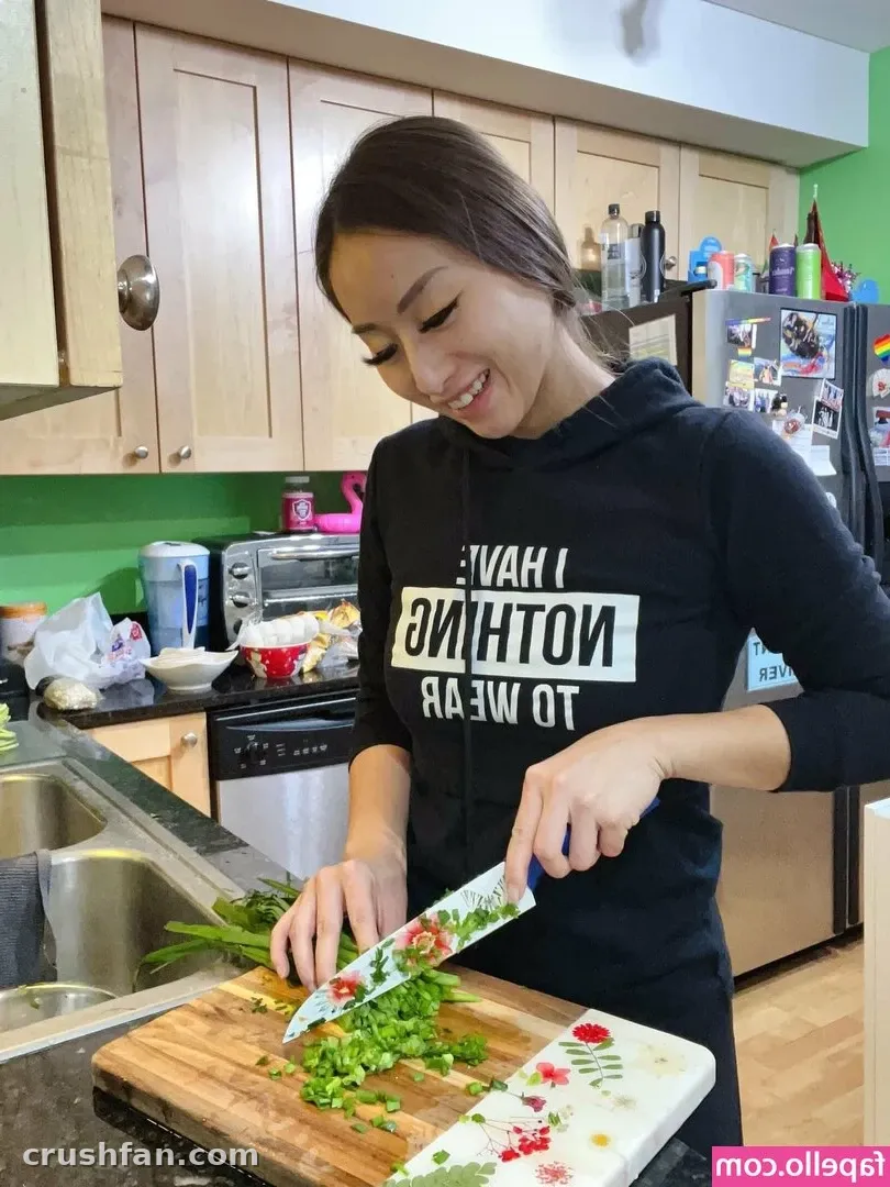 Smiling woman in a black hoodie chopping green herbs on a wooden board in a bright kitchen
