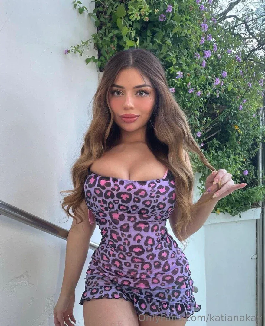 Katiana Kay in a purple leopard print dress standing outdoors by a white wall and green plants