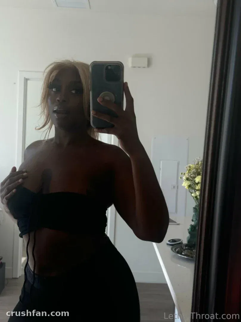 Lethal Throat taking a mirror selfie in a dimly lit room wearing a dark strapless top.