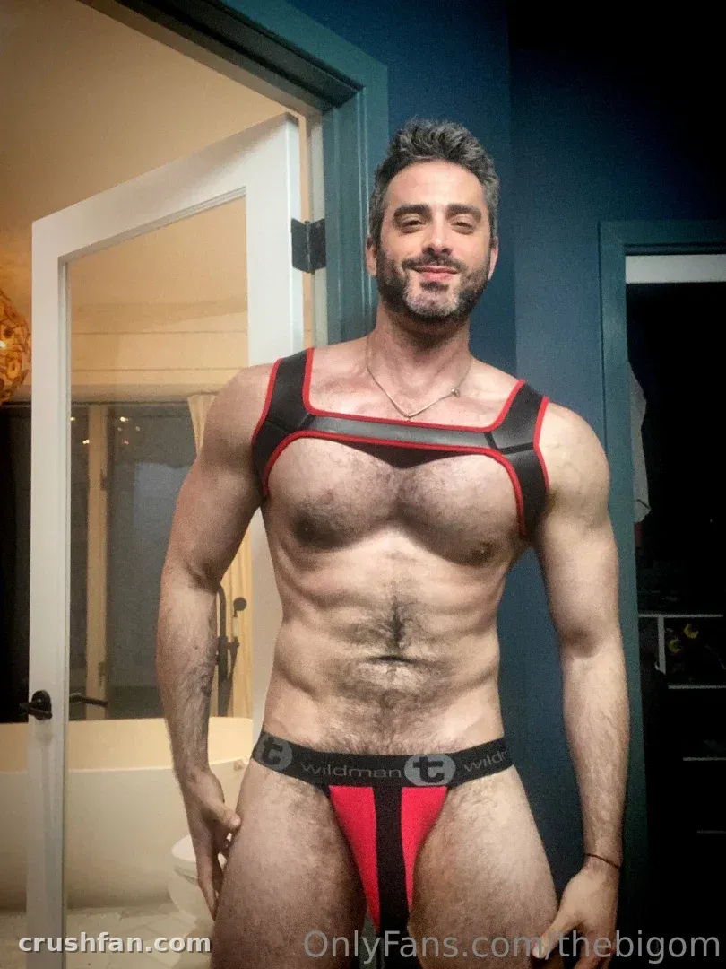 The Big Om with Kae in a red and black harness and jockstrap indoors
