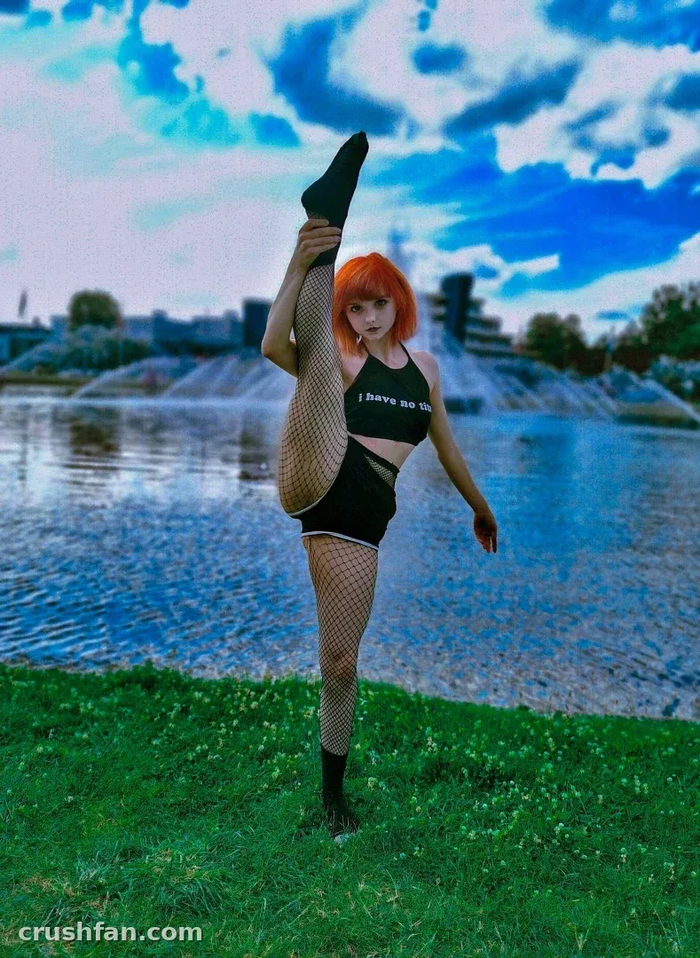 Creator in black top and fishnets holding a high leg lift on grass beside water.