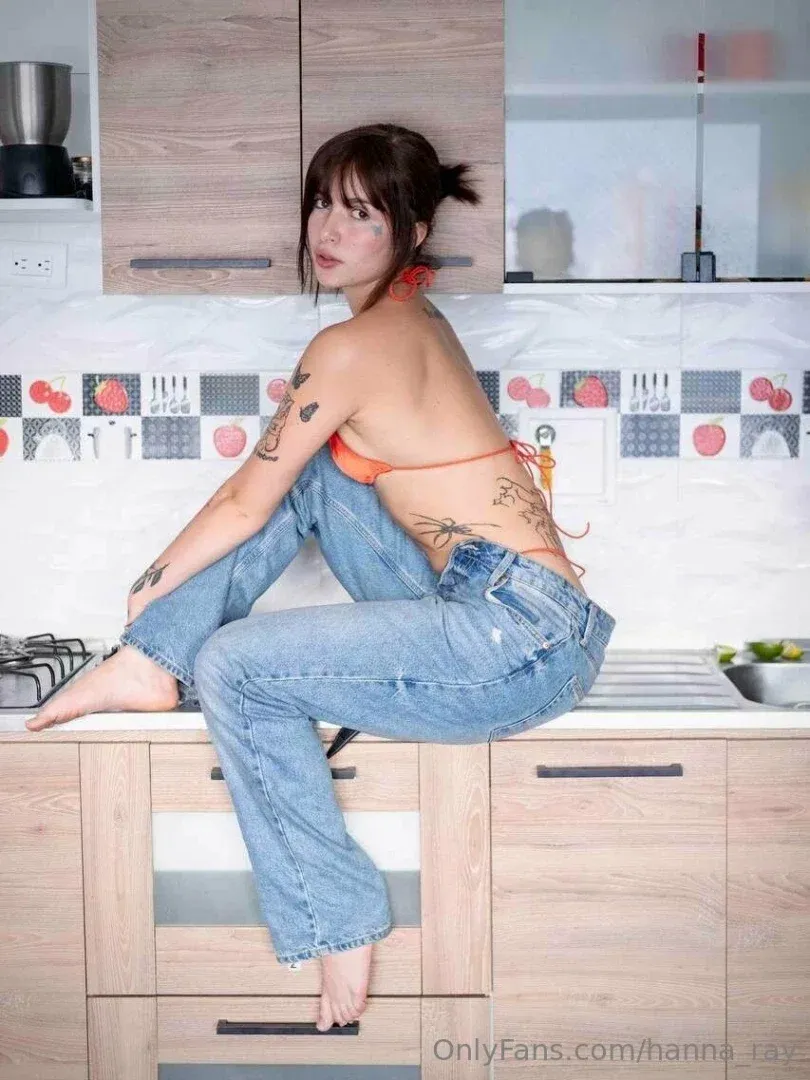 Woman in orange bikini and blue jeans sitting on a kitchen counter, looking back at the camera.
