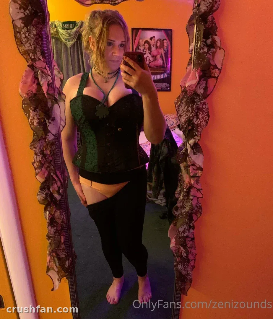 Zeni Zounds in a green and black corset taking a mirror selfie in an orange-lit room.