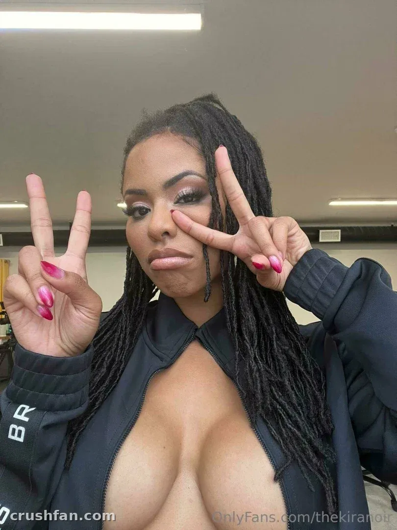 Kira Noir con una chaqueta oscura haciendo signos de paz con uñas rosas en interiores.