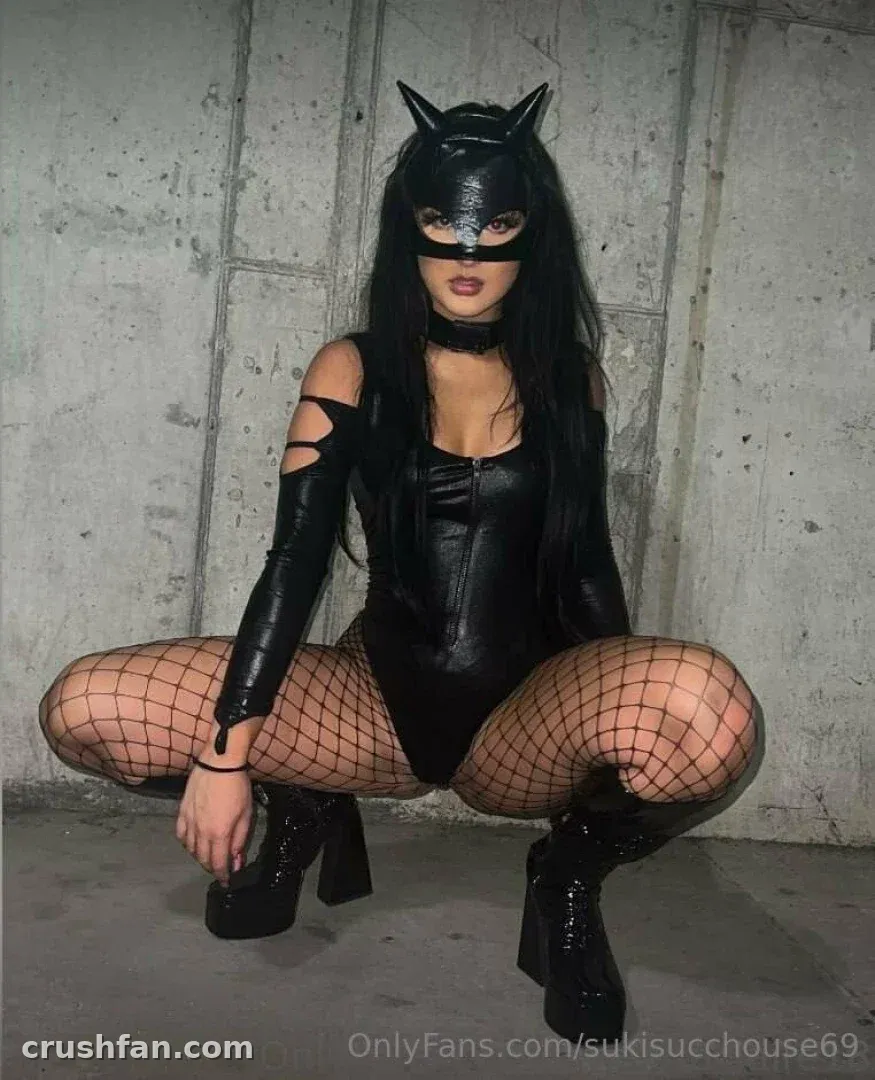 Suki in einer schwarzen Katzenmaske und einem Leder-Bodysuit, geduckt vor einer Betonwand