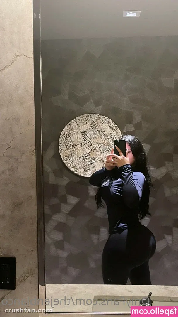 Jen in a black top and leggings taking a mirror selfie in a bathroom with a round wall decoration.