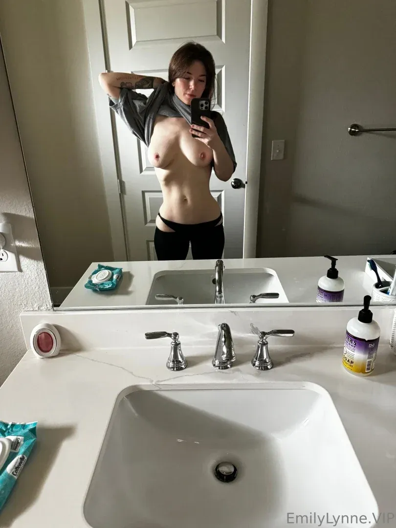 Emily Lynne tirando uma selfie no espelho em um banheiro, usando uma camisa cinza aberta