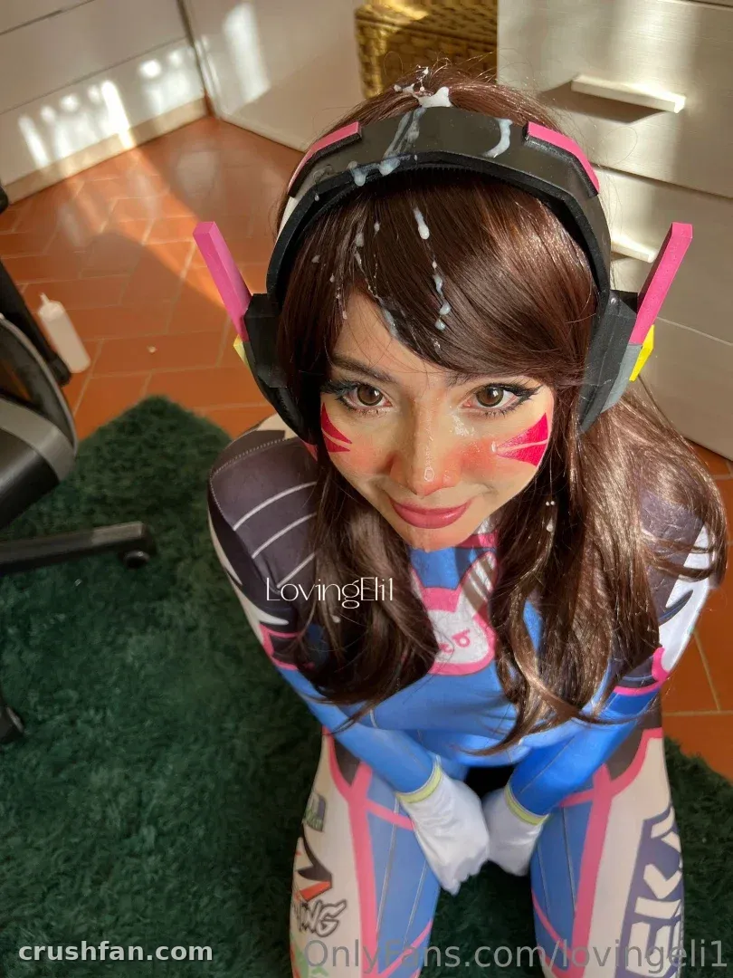 Eli em um traje de cosplay azul e rosa com pintura facial e um headset, inclinando-se em direção à câmera