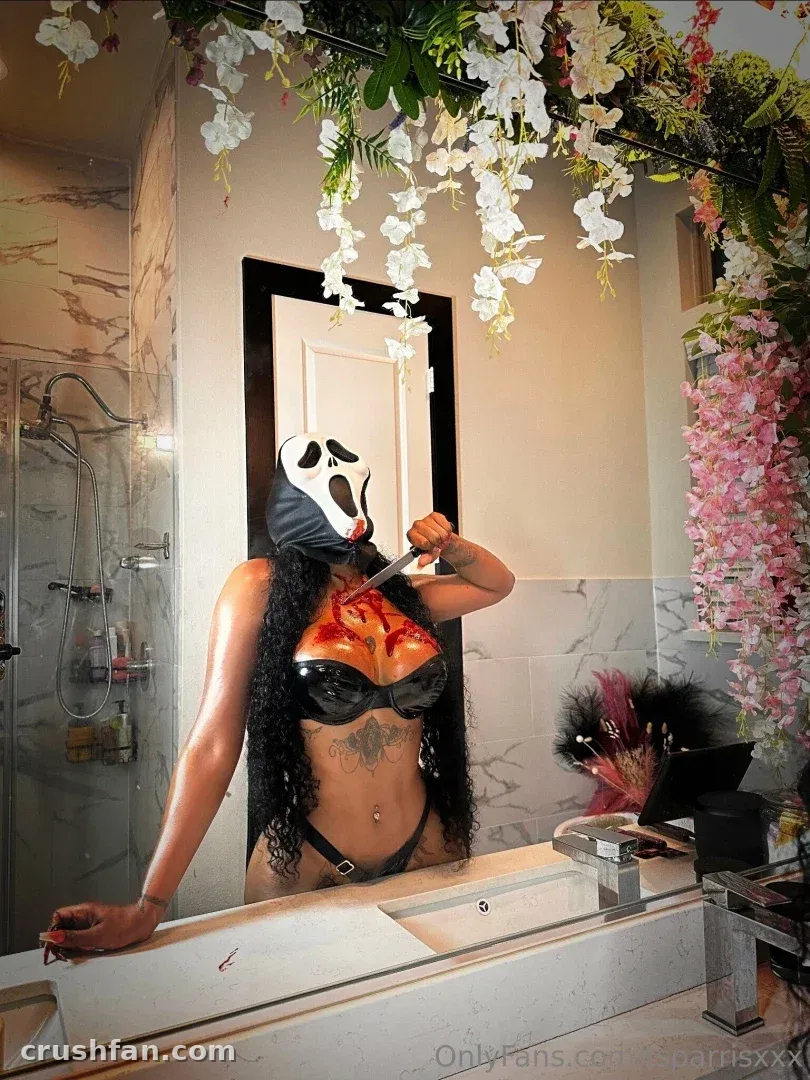Creator in a Ghostface mask and black bra taking a mirror selfie in a flower-decorated bathroom.