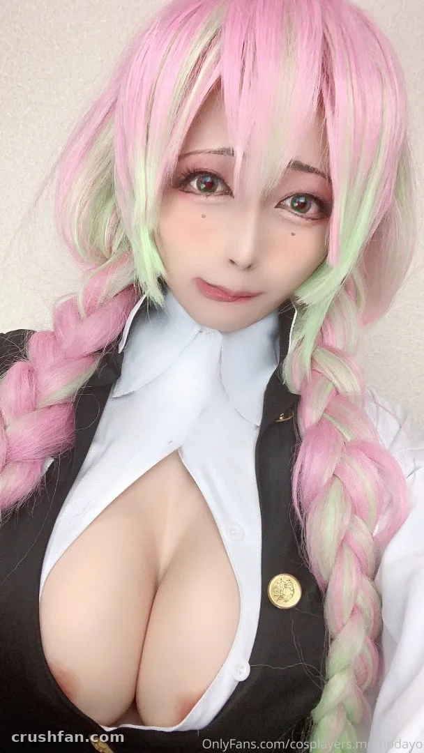 Cosplayer in a pink and green braided wig, white collar and dark vest, facing the camera.