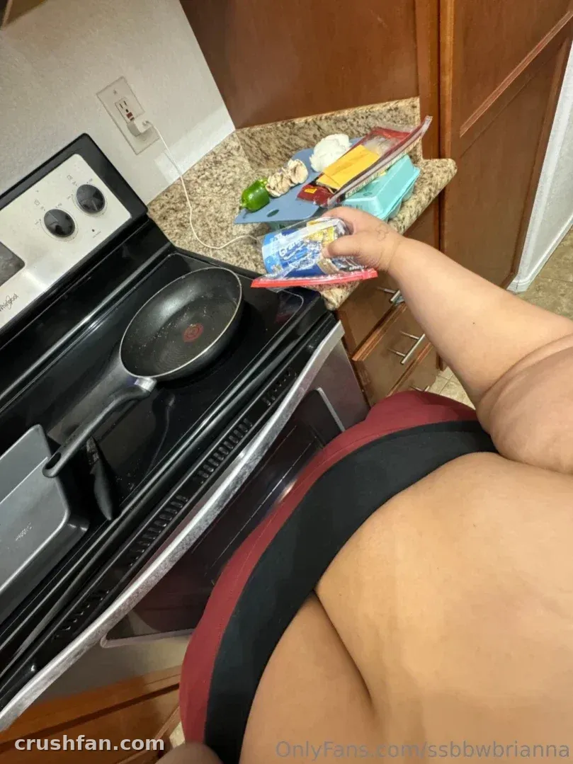 Creadora con un sujetador deportivo oscuro de pie junto a una estufa con una sartén y comida en la encimera.