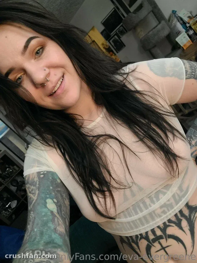 Eva Evergreene sorrindo com um top branco transparente e braços tatuados em ambiente interno