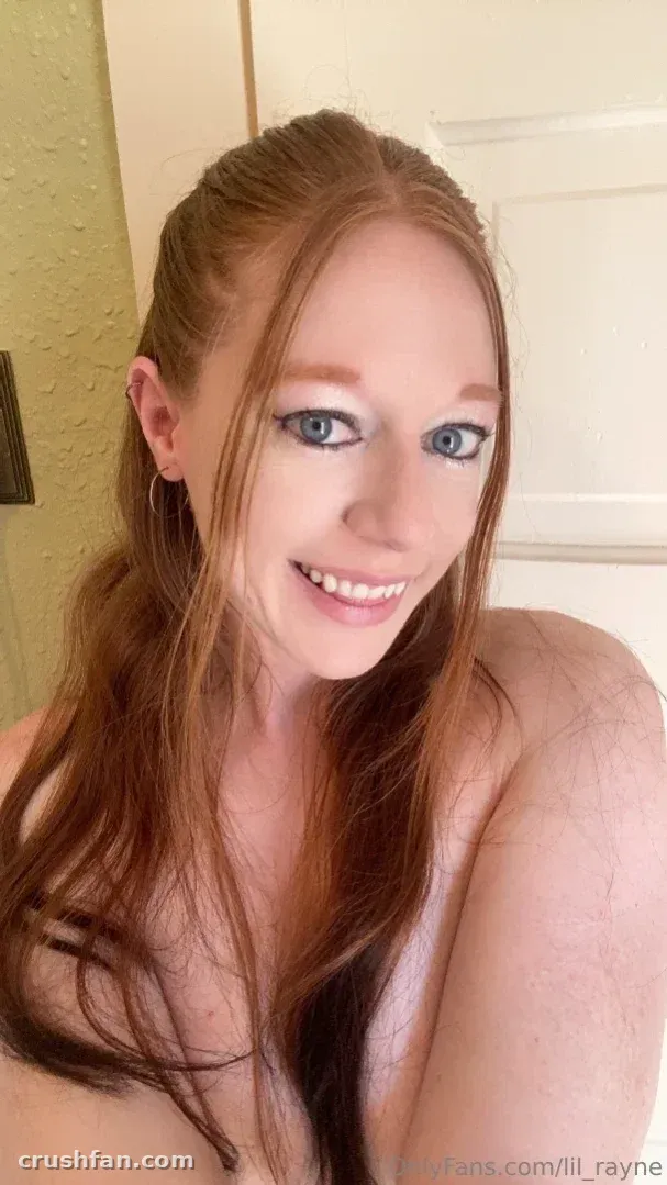 Close-up selfie of a smiling woman with long auburn hair in soft indoor light