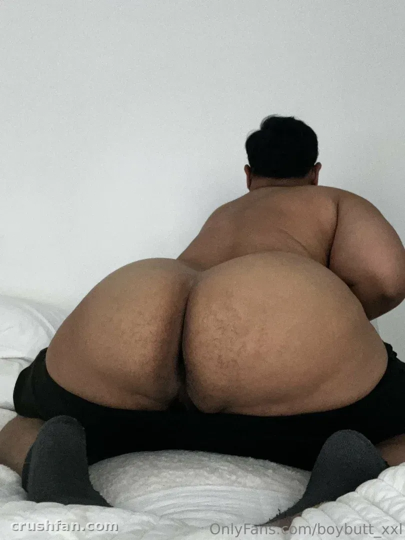 BoyButtXXL arrodillado en una cama blanca con pantalones cortos negros contra una pared blanca lisa