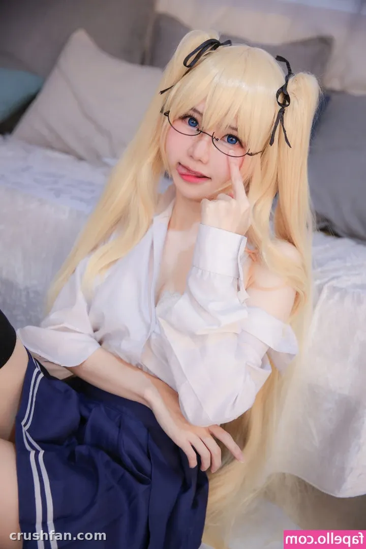 Sally Dorasnow in a blonde twin-tail wig and glasses, wearing a white blouse and navy skirt on a bed