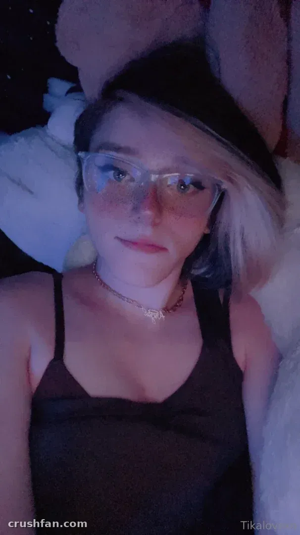 Tika tirando uma selfie de perto em ambiente interno sob luz azul baixa, usando óculos e uma regata escura.