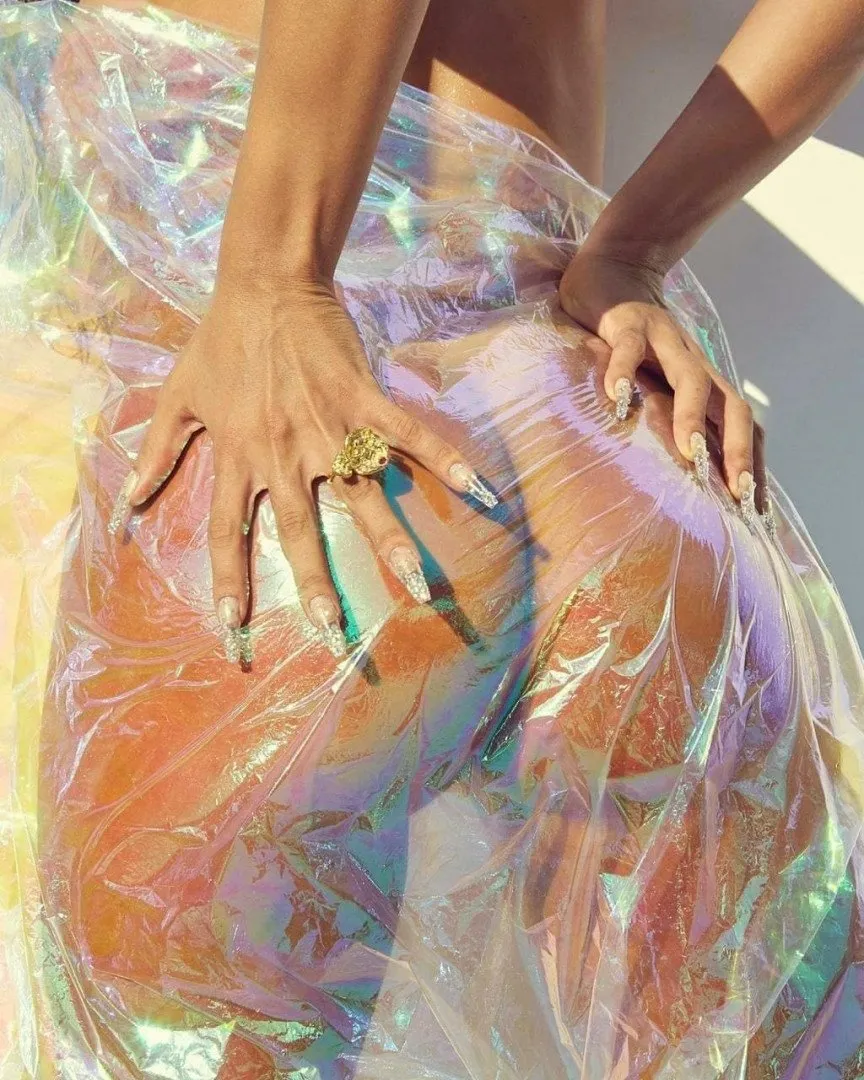 Anitta avec des mains bijoutées posées sur un tissu transparent iridescent sous une lumière de studio vive.