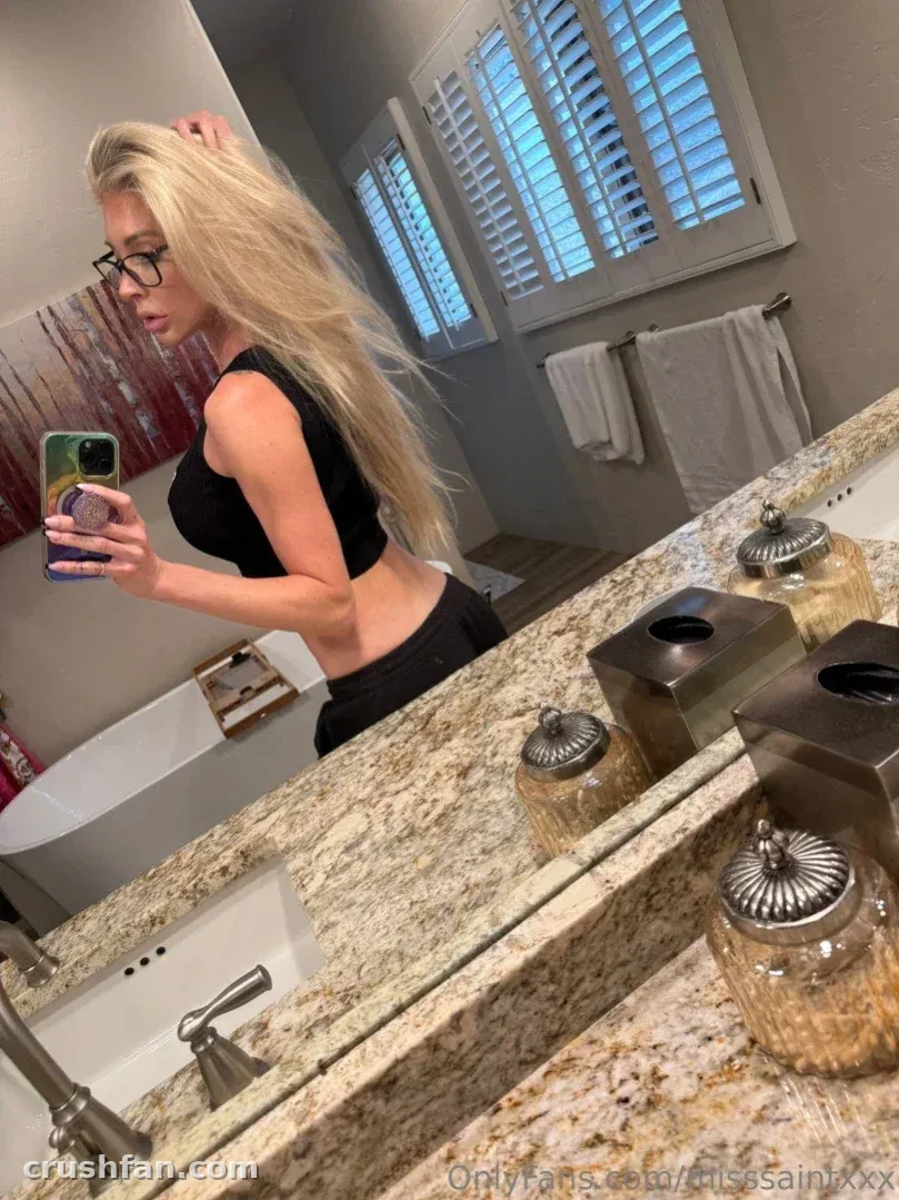 Mujer rubia con blusa negra haciéndose una selfie frente al espejo en un baño luminoso con encimeras de granito