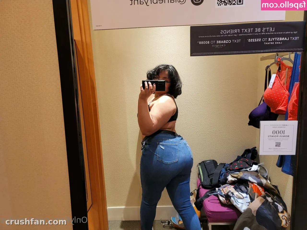 Criadora com uma blusa escura e calça jeans azul tirando uma selfie no espelho em um provador com uma arara de lingerie atrás dela.