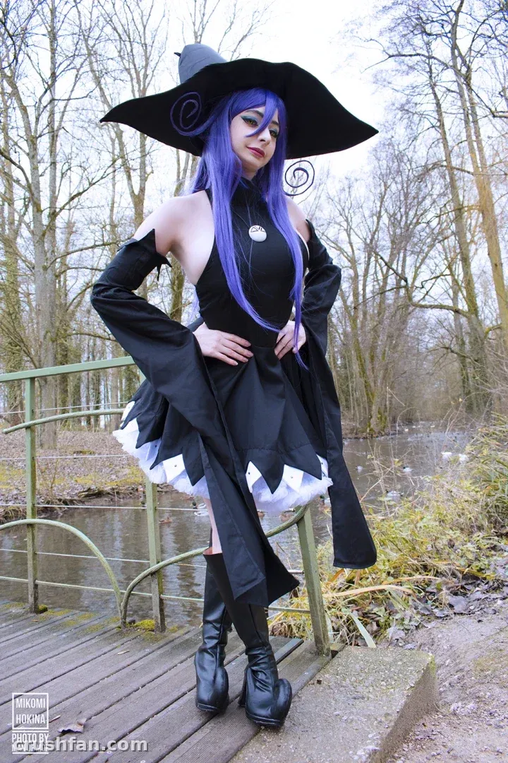 Cosplayer com uma fantasia de bruxa preta e chapéu de aba larga em pé em uma ponte de madeira perto de um lago
