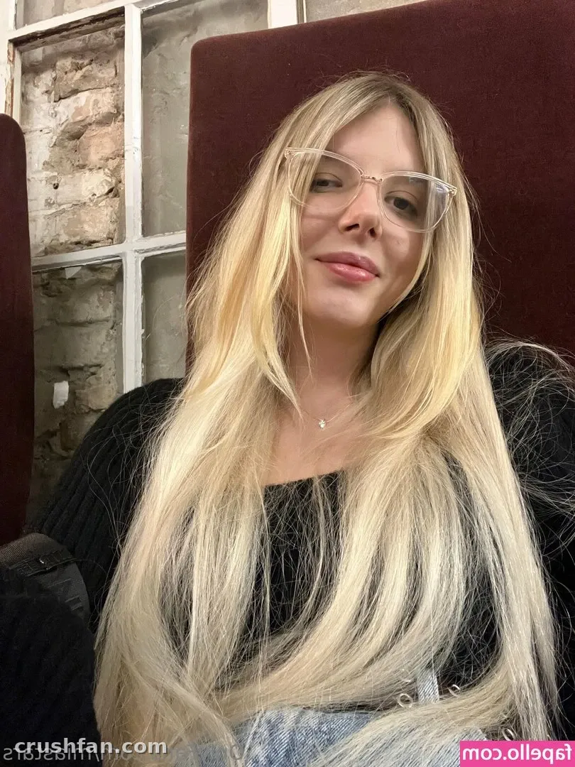 Mia de óculos e jardineira jeans sentada em ambiente interno contra um encosto vinho
