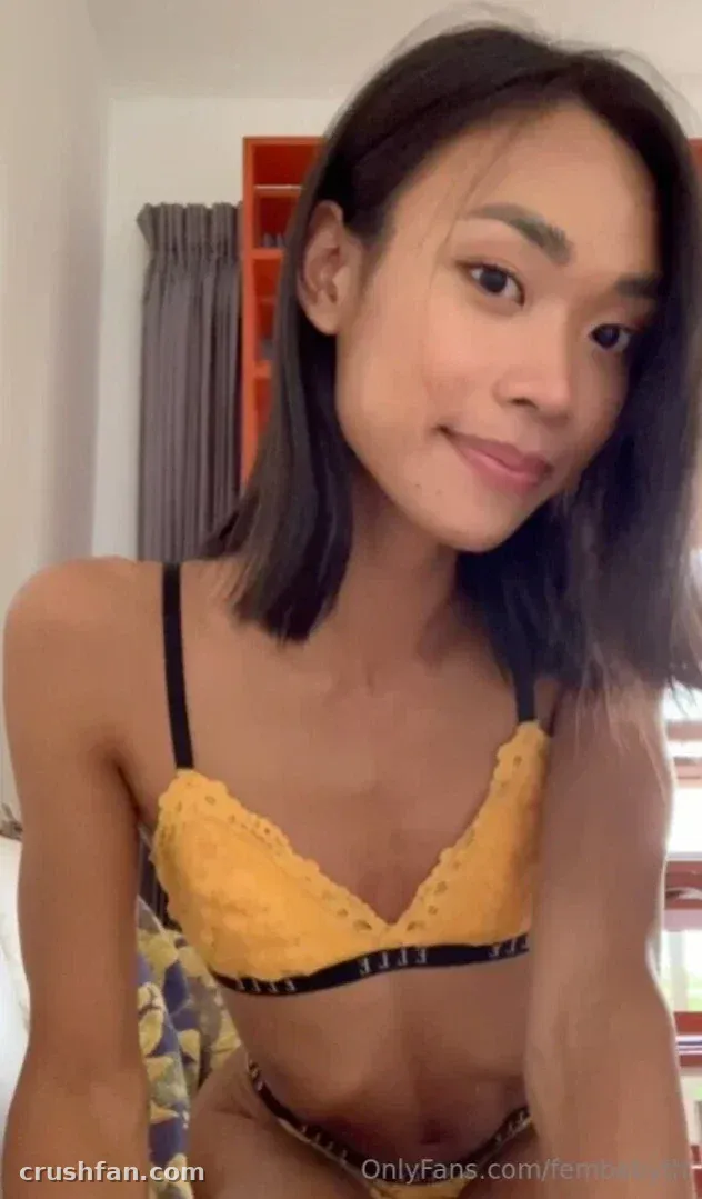 Femby con un bralette de encaje amarillo posando en interiores con una cortina gris y una estantería roja detrás de ella.
