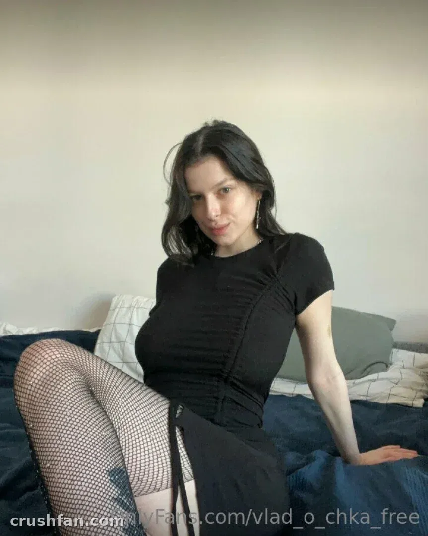 Vladochka com uma blusa e saia pretas com meia-calça de rede sentada em uma cama
