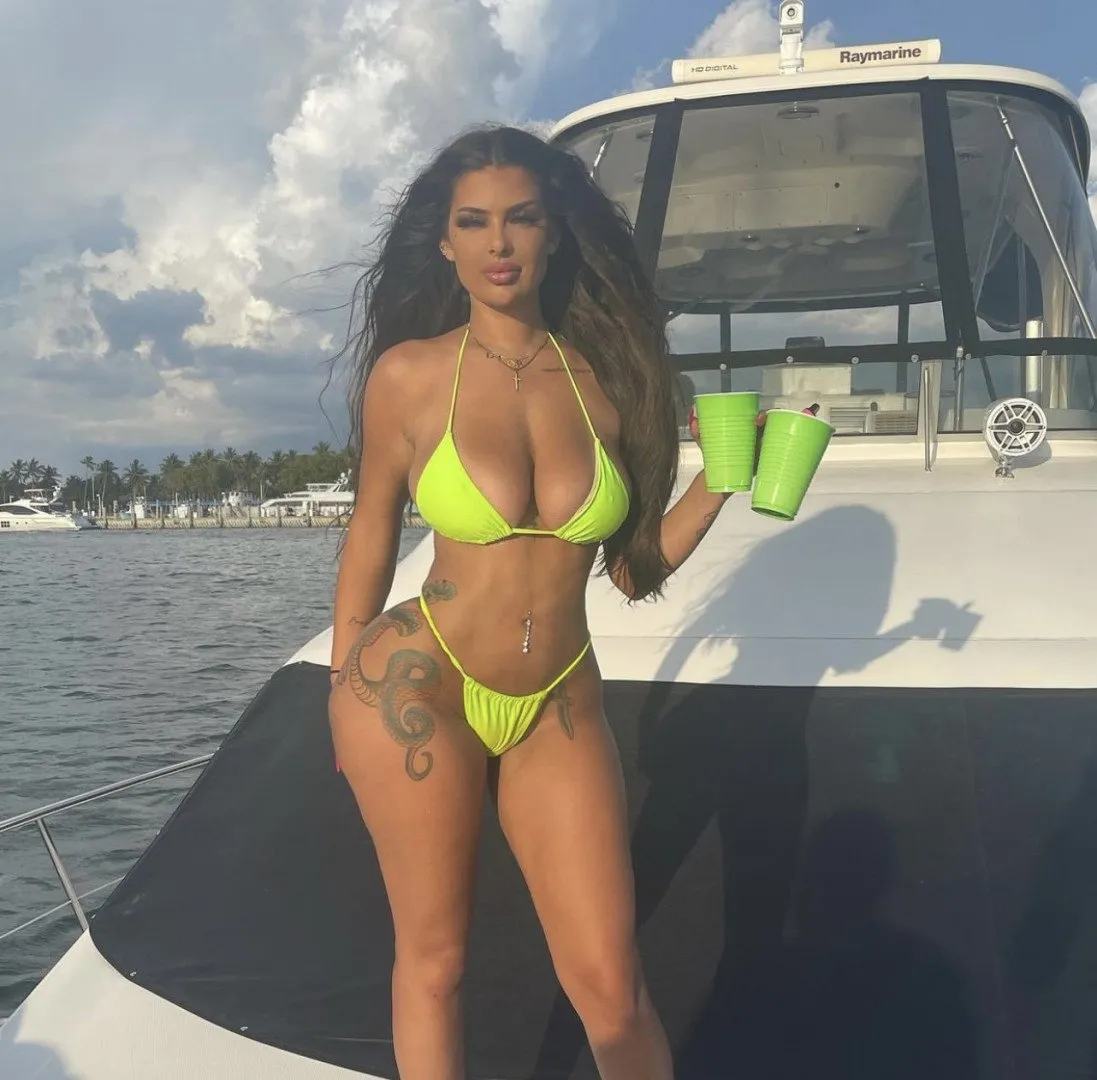 Der griechische Freak in einem neon-grünen Bikini, der zwei grüne Becher auf einem Boot hält.