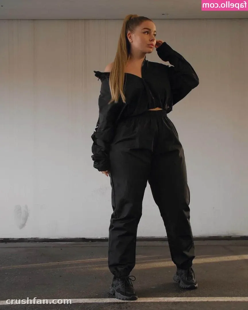 Creadora con un top de hombros descubiertos y pantalones totalmente negros de pie frente a una pared lisa