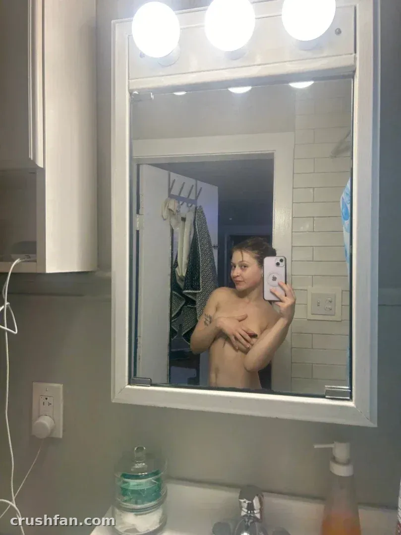 Kali taking a mirror selfie in a bright bathroom with white tiles and round bulbs.