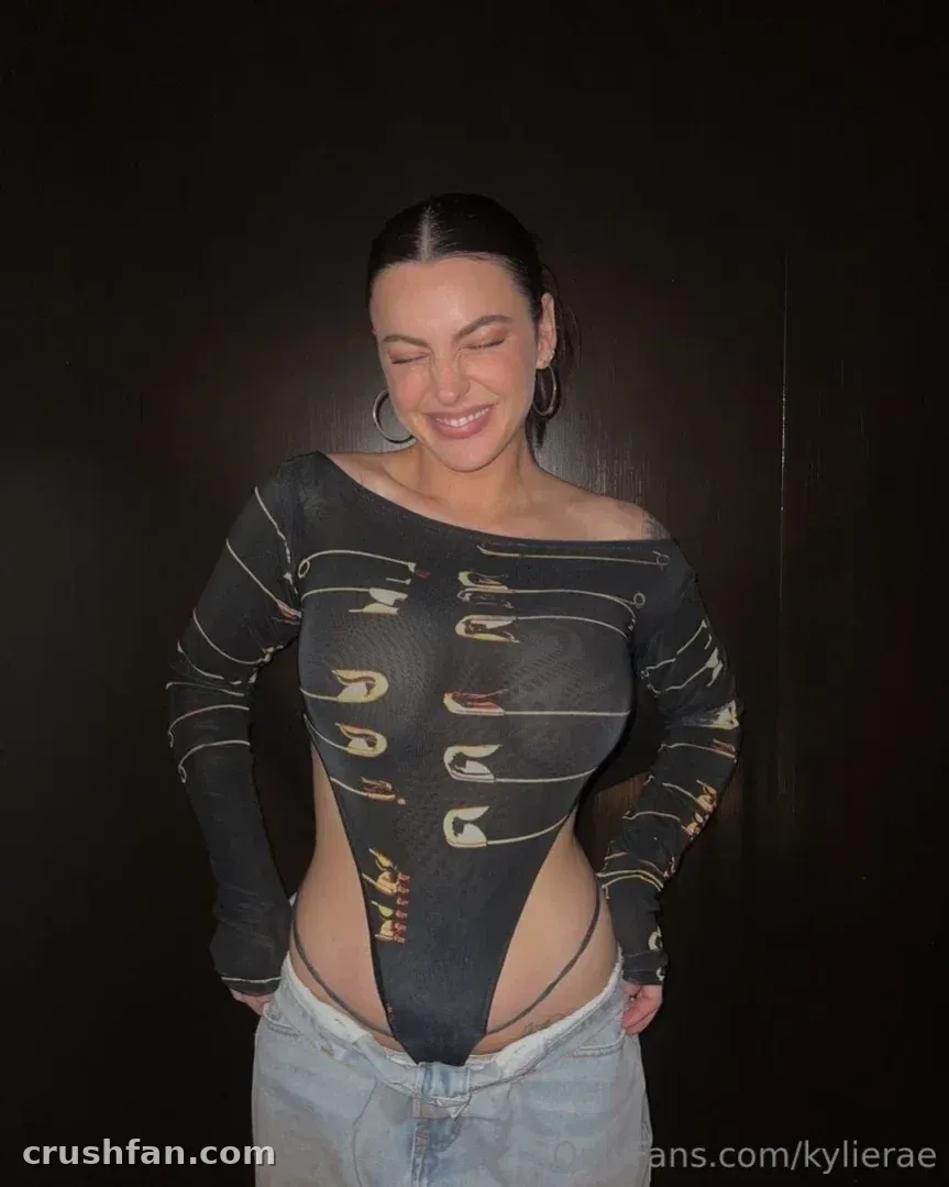 Kylie Rae souriant dans un body à motif graphique et un jean clair contre un mur sombre