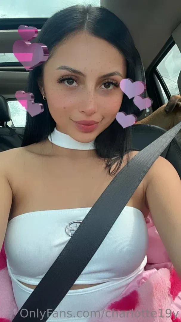 Charlotte avec un haut blanc et un choker prenant un selfie en voiture avec des autocollants en forme de cœur rose