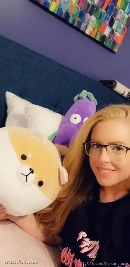 Vicktoria Tacos in glasses holding a plush dog toy on a bed with pillows and colourful wall art