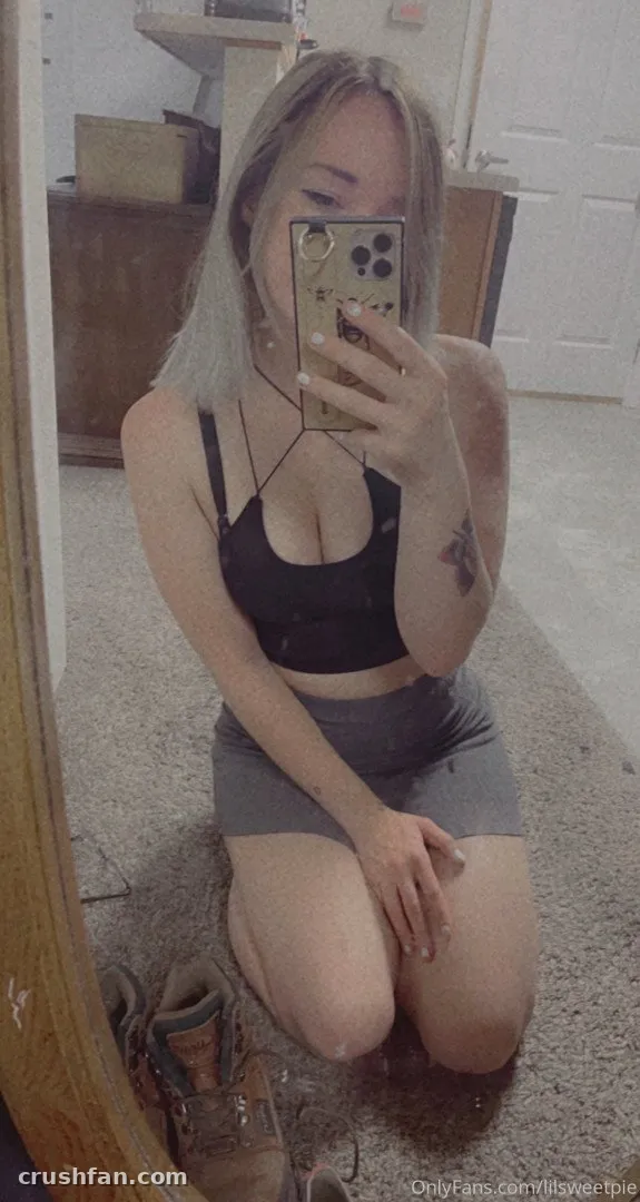 Amelia kneeling on a carpeted floor taking a mirror selfie in a black crop top and grey skirt.