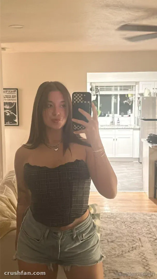 Lexi com um top xadrez e shorts jeans tirando uma selfie no espelho em ambiente interno.