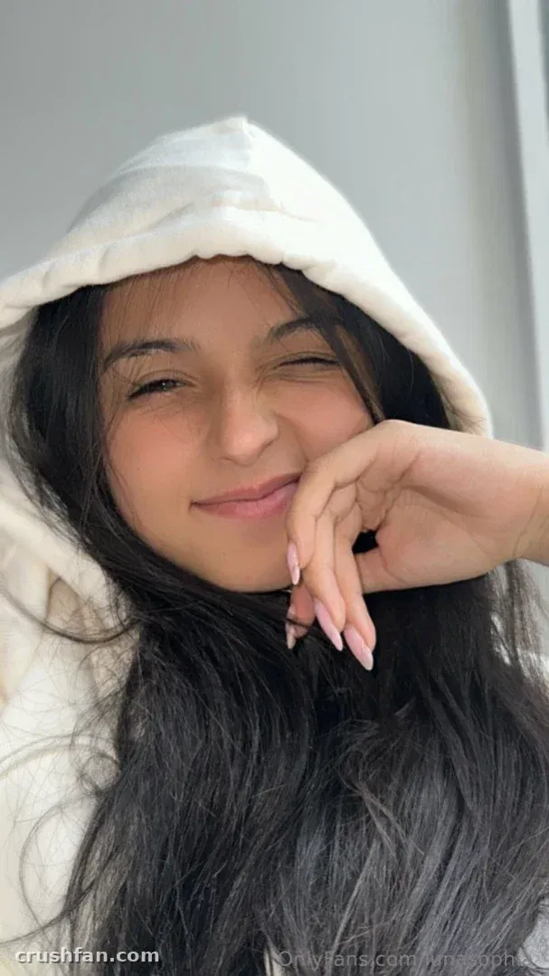 Luna Sophia winking in a white hooded top against a light grey wall