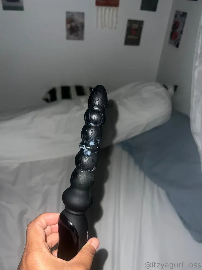 Una mano sostiene un juguete texturizado negro brillante en un dormitorio con iluminación suave y ropa de cama blanca.