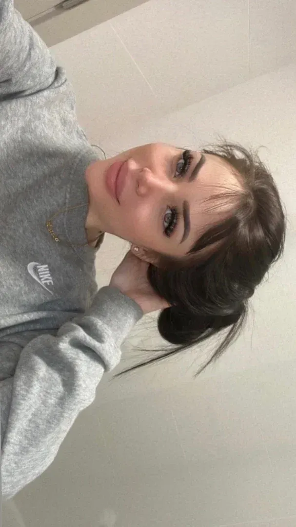 Estela in einem grauen Sweatshirt, die drinnen vor einer hellen Wand ein Nahaufnahme-Selfie macht.
