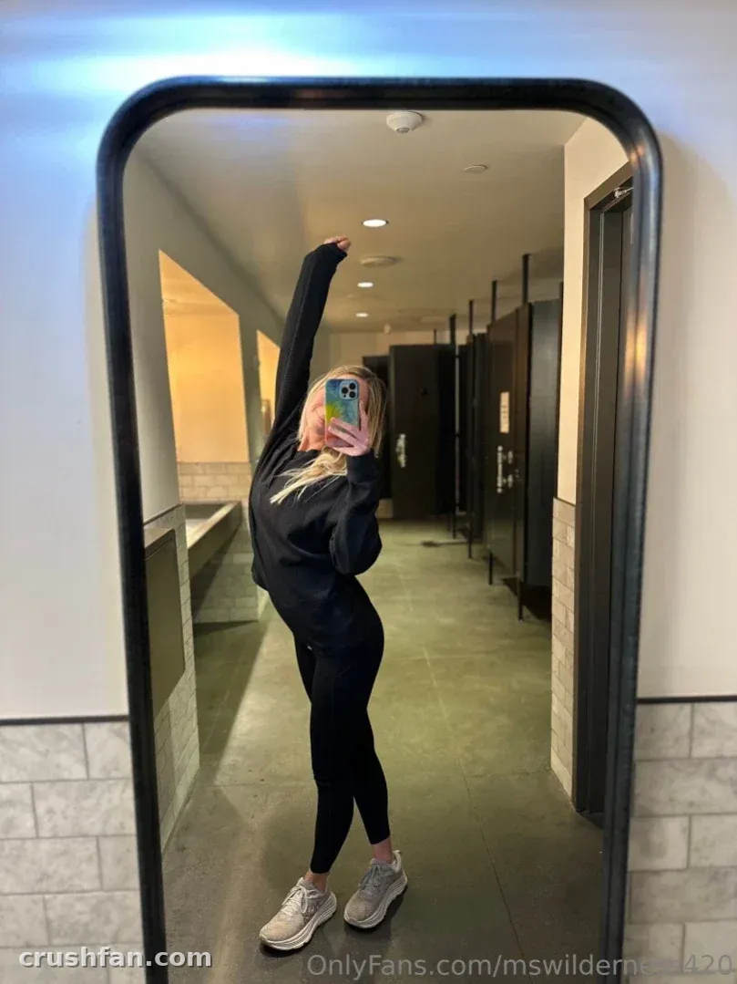 Ms Wilderness takes a mirror selfie in a hallway wearing a black top and leggings.