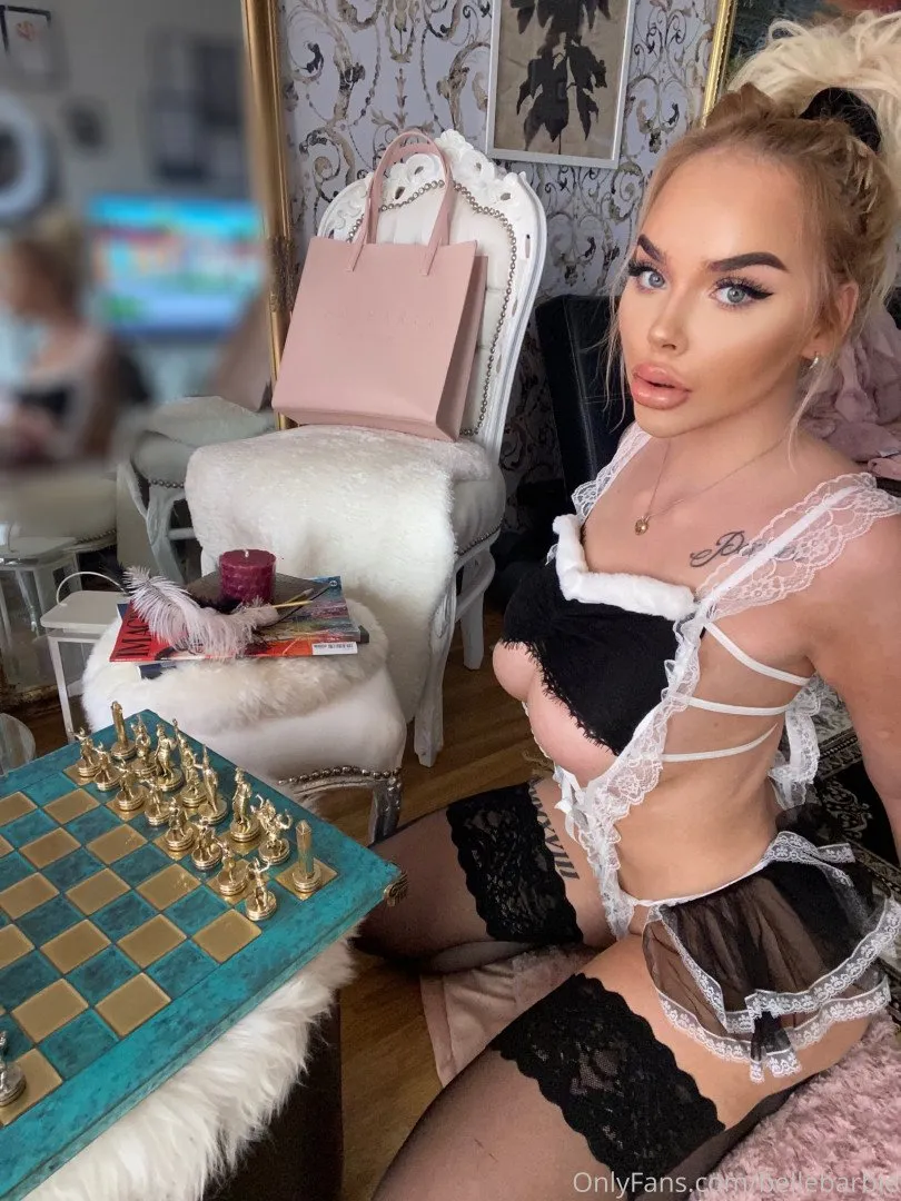 Criadora com lingerie de renda preta e branca sentada ao lado de um tabuleiro de xadrez verde-azulado em uma sala aconchegante.