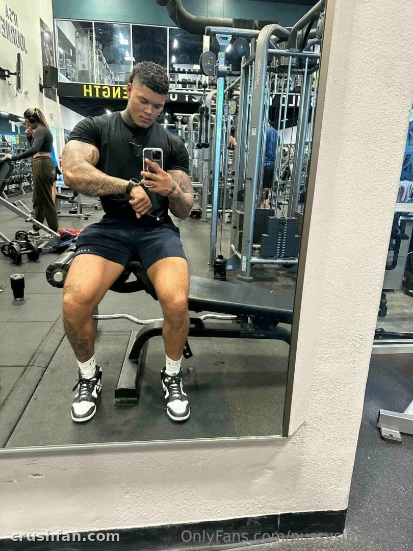 DOMINATOR en t-shirt noir et short prenant un selfie dans le miroir d'une salle de sport.