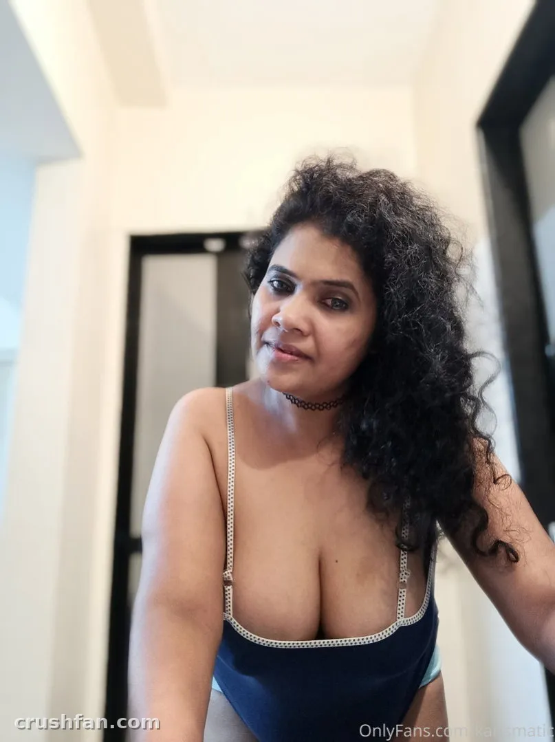Karishma dans un haut bleu marine à bretelles chaîne et ras-du-cou, debout en intérieur avec des cheveux bouclés.