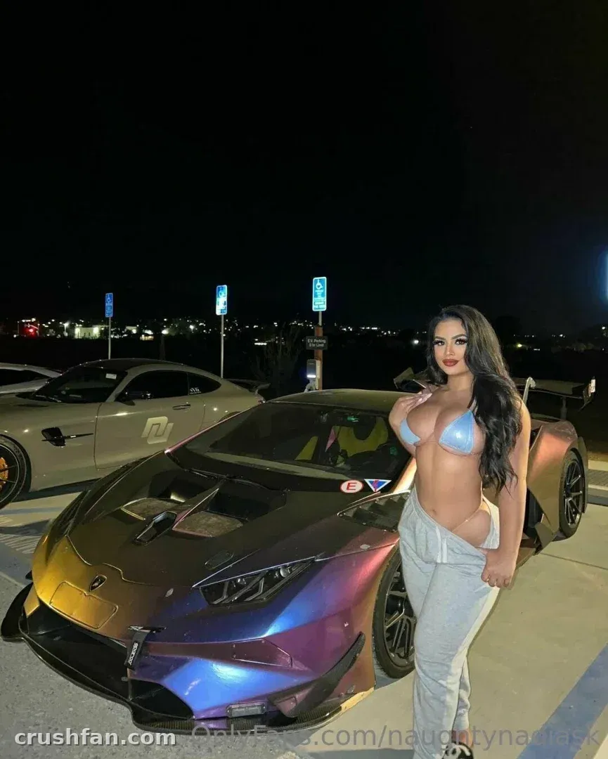 Nadia con un top de bikini azul y pantalones grises de pie junto a un deportivo con pintura camaleónica de noche.