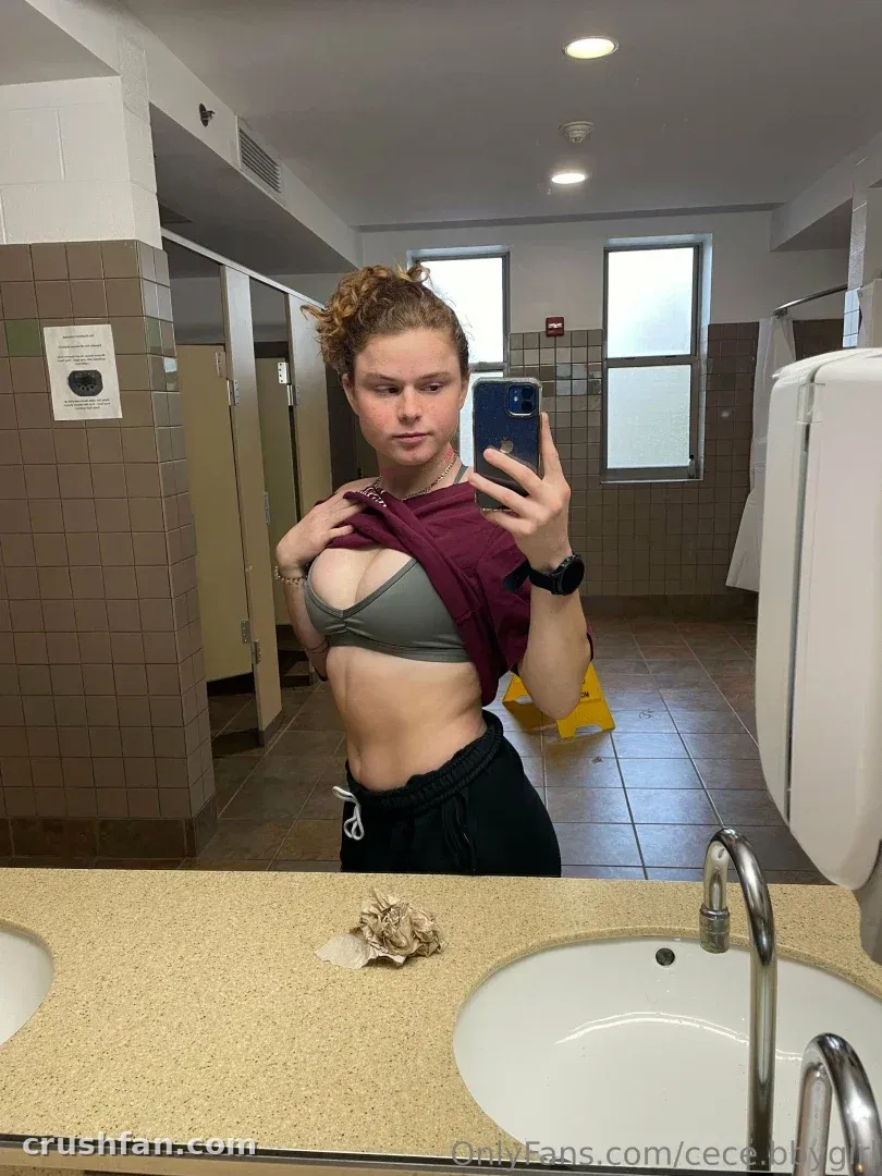 Selfie no espelho de uma mulher com blusa vinho e top esportivo cinza dentro de um banheiro azulejado