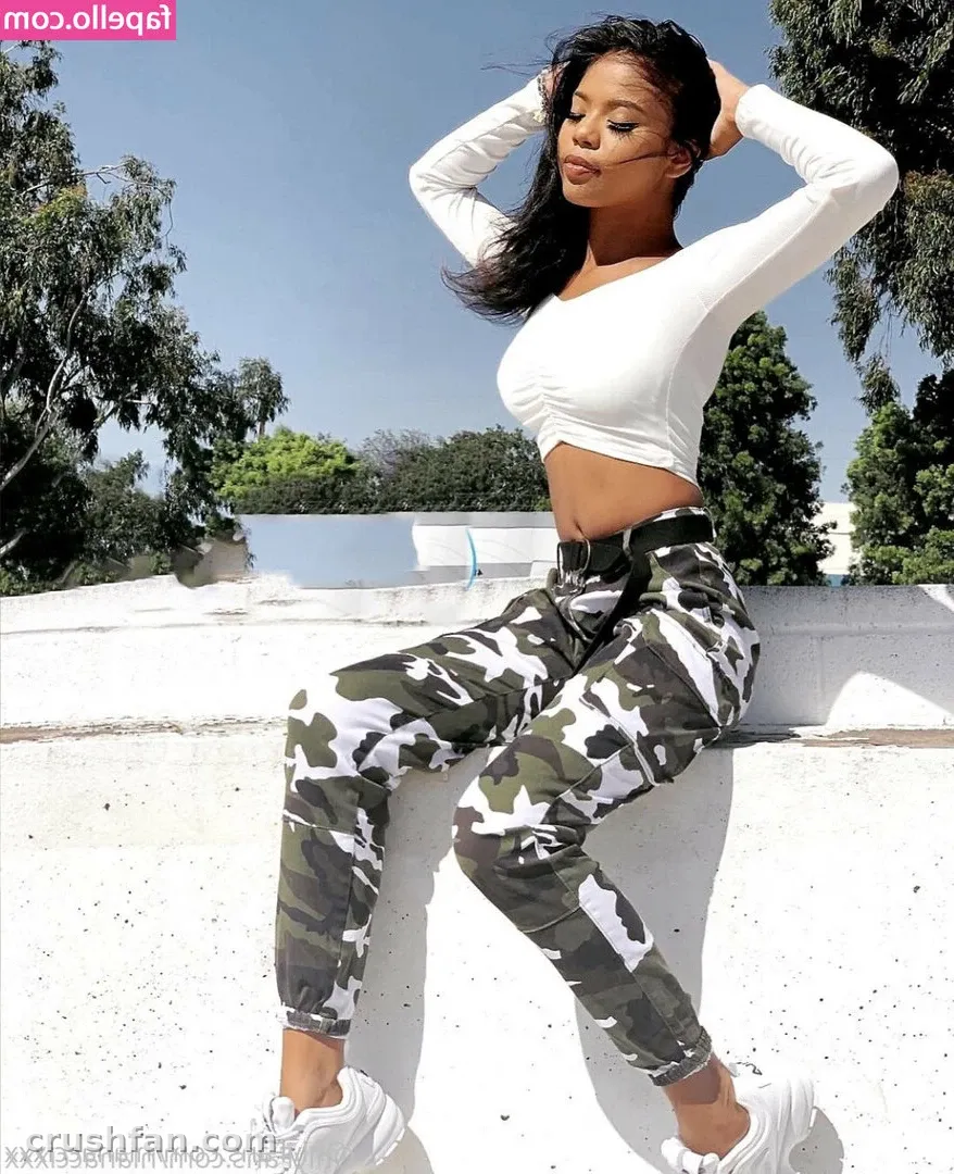 Nia Nacci com um top cropped branco e calça de camuflagem sentada em um muro branco ao ar livre