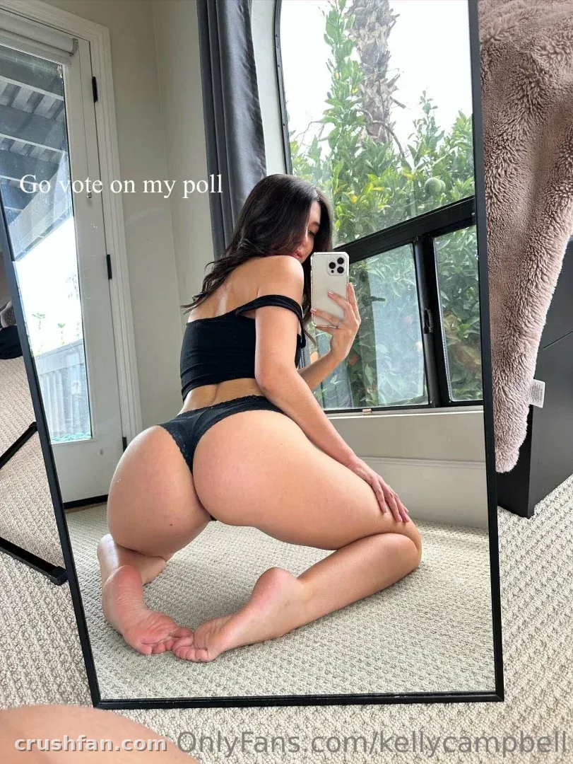 Kelly Cams kauert auf einem Teppich für ein Spiegel-Selfie in einem hellen Raum mit Fenstern und Pflanzen.