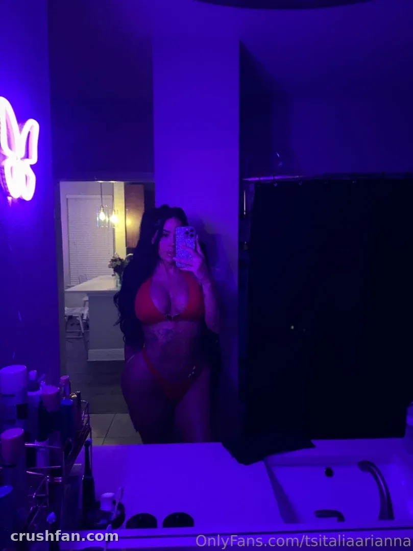Italia Arianna macht ein Spiegel-Selfie in roter Unterwäsche unter violettem Neonlicht in einem Badezimmer.