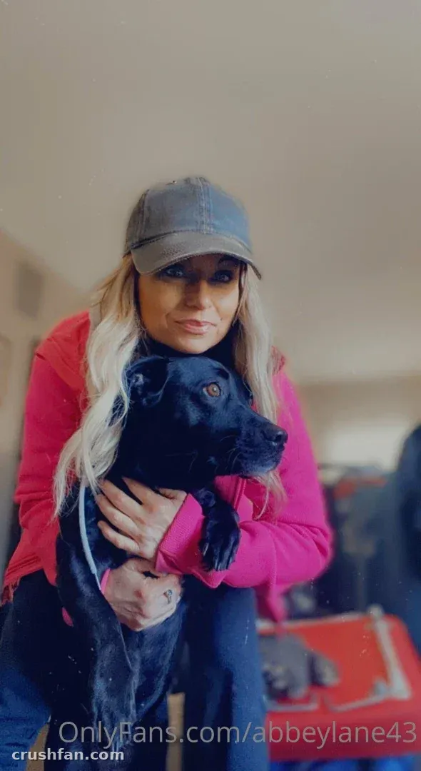 Abbey Lane com um top rosa e boné cinza segurando um cachorro preto em ambiente interno
