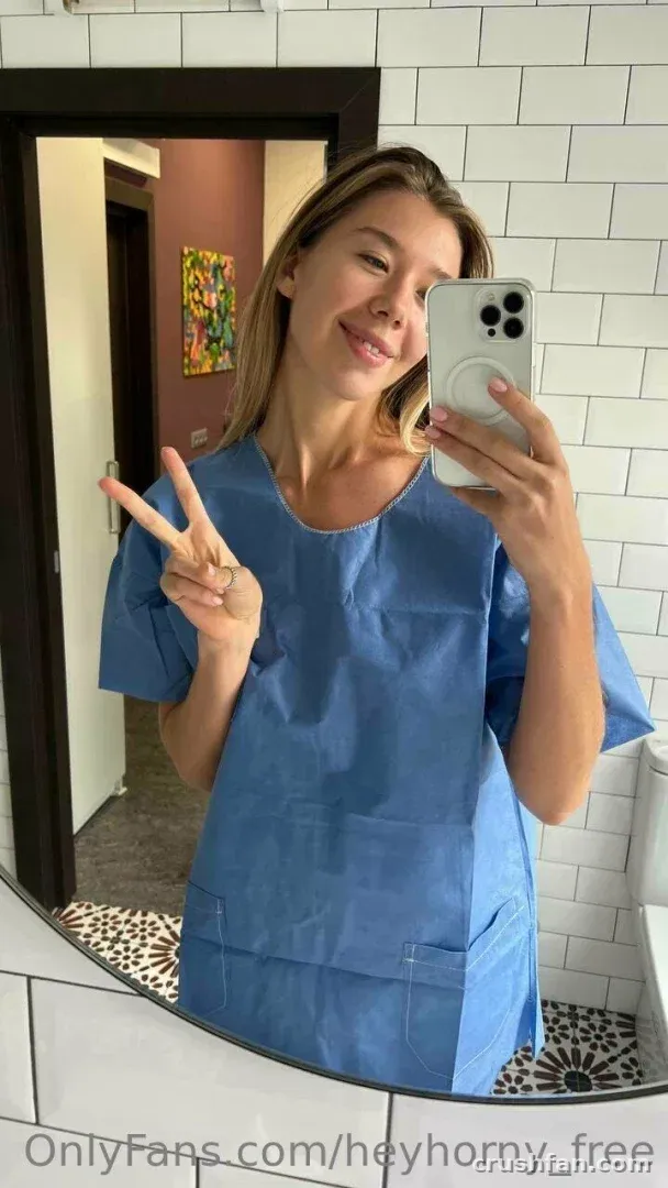 Creator in blue scrubs taking a mirror selfie with a peace sign in a tiled room.